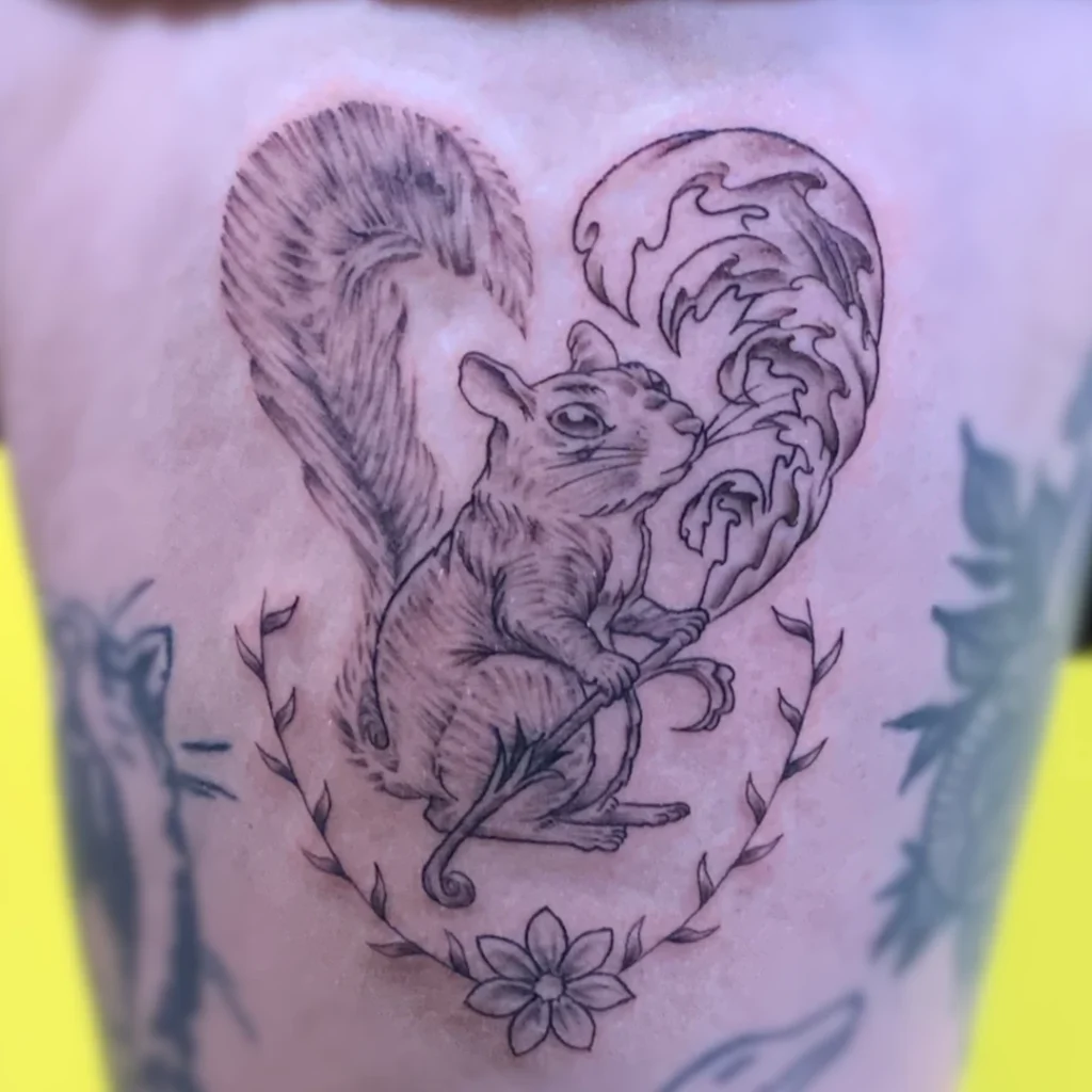 A thigh tattoo of a squirrel holding a leaf, the tail and leaf form the shape of a heart. Rendered in black and grey.