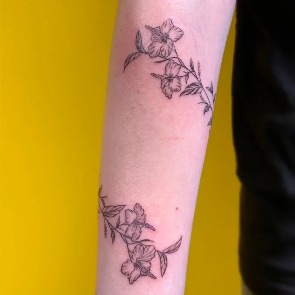 A fine line forearm tattoo of hibiscus flowers growing on a vine that wraps around the arm twice.