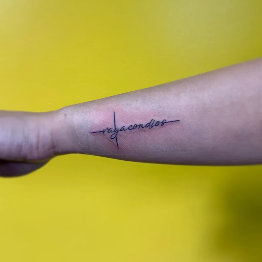 Wrist tattoo of a cross with "vaya con dios" written in the center.